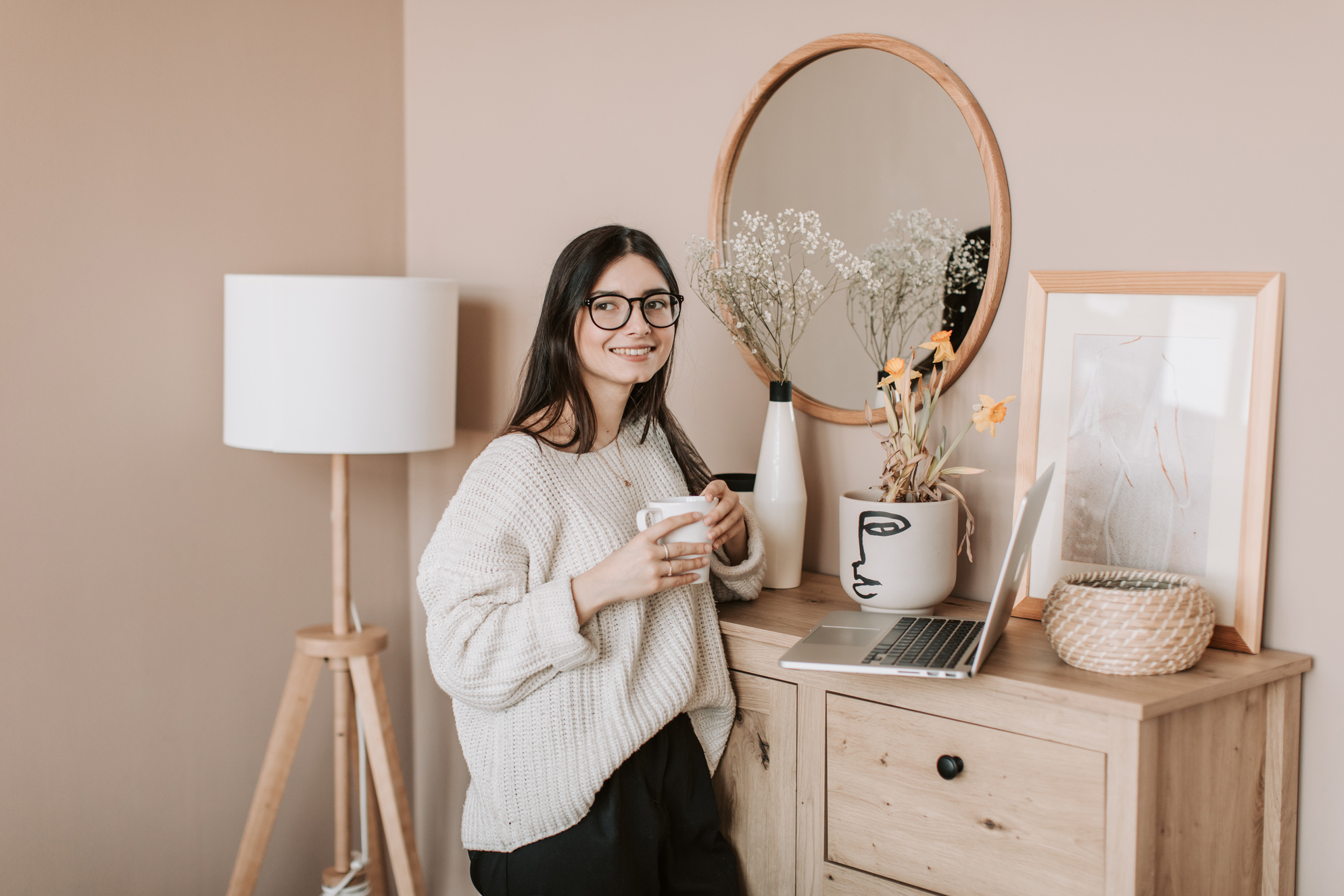 Young woman with cup of drink and laptop near dresser